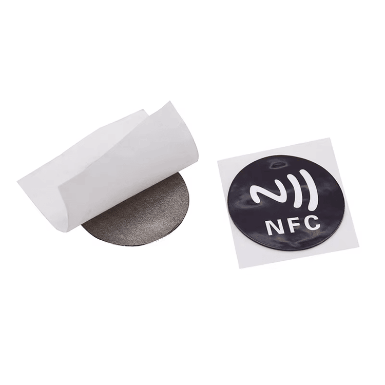 Fotografija proizvoda prikazuje okruglu PVC RFID oznaku za metal s NFC licem pored odlepljenih slojeva poleđine za identifikaciju na metalu.
