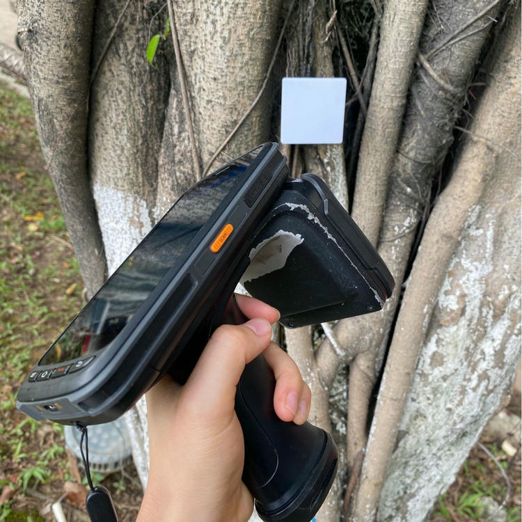 Slika prikazuje ručni RFID čitač koji skenira belu oznaku NAIL06-7 postavljenu na drvo radi terenskog ispitivanja identifikacije.