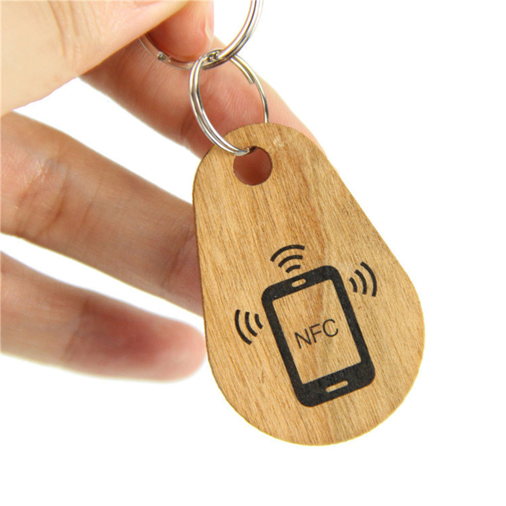 Ruka drži drveni RFID privitak za ključeve u obliku suze sa štampanom ikonom NFC telefona i radio talasima za brzu kontrolu pristupa dodirom.