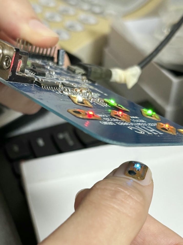 Svetleća FLED1006-1 NFC LED oznaka prikazana je na jagodici prsta blizu elektronske testne ploče sa aktivnim jarkim plavim svetlom.