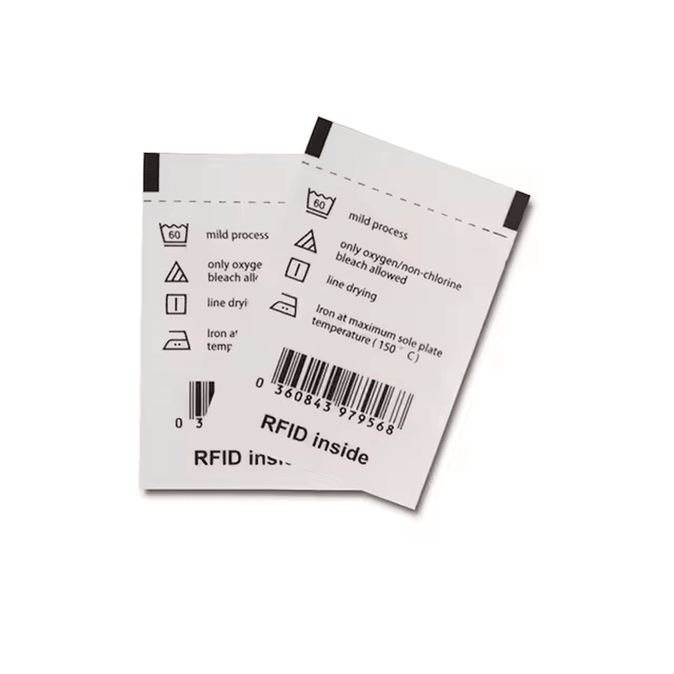 Dve bele RFID tekstilne etikete za pranje prikazane su preklopljene sa ikonama za pranje i štampanim barkodom za upravljanje odećom.