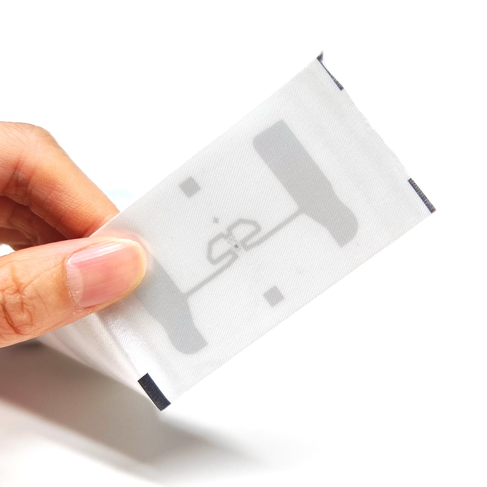 Ruka drži kompaktnu RFID etiketu za odeću blizu kamere kako bi se istakli savitljiva tkana površina i vidljiv raspored antene.
