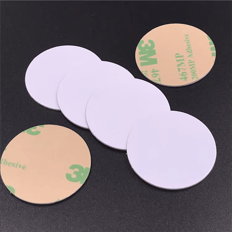 Prazni beli RFID disk tagovi prikazani su sa lepljivim podlogama na tamnoj pozadini koja ističe montažu lepljenjem i odvajanjem.