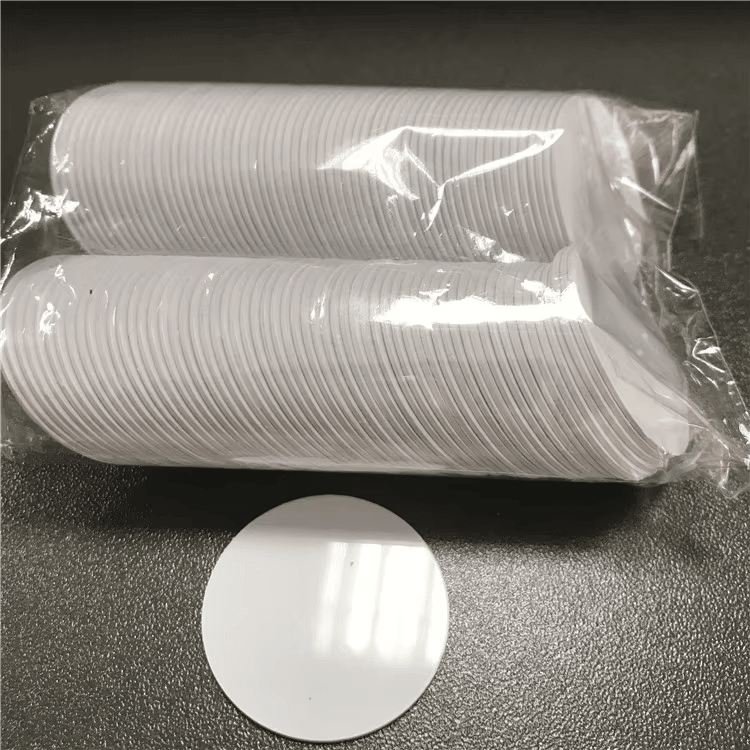 Dve zapakovane hrpe belih RFID disk tagova nalaze se iznad jednog praznog diska i prikazuju kako se coin tagovi isporučuju u velikim količinama.