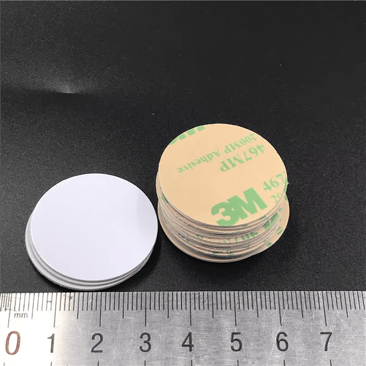 Prednja i zadnja hrpa RFID disk tagova prikazane su pored lenjira koji upoređuje belu prednju stranu sa stranom lepljive podloge.