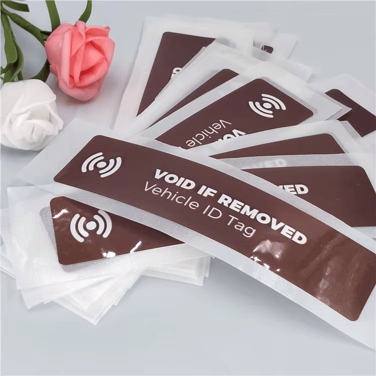 Sjajne RFID etikete za vetrobran koje pokazuju pokušaj uklanjanja preklapaju se na stolu uz prikaz zakrivljenih krajeva i teksta VOID IF REMOVED za zaštitu od krađe.