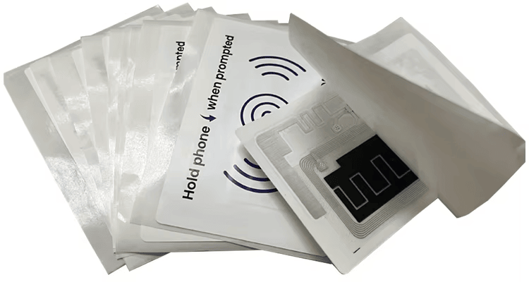 Ova slika prikazuje više samolepljivih temperaturnih RFID nalepnica naslaganih jedna na drugu, sa jednom otvorenom nalepnicom koja otkriva pametni senzorski inlej.